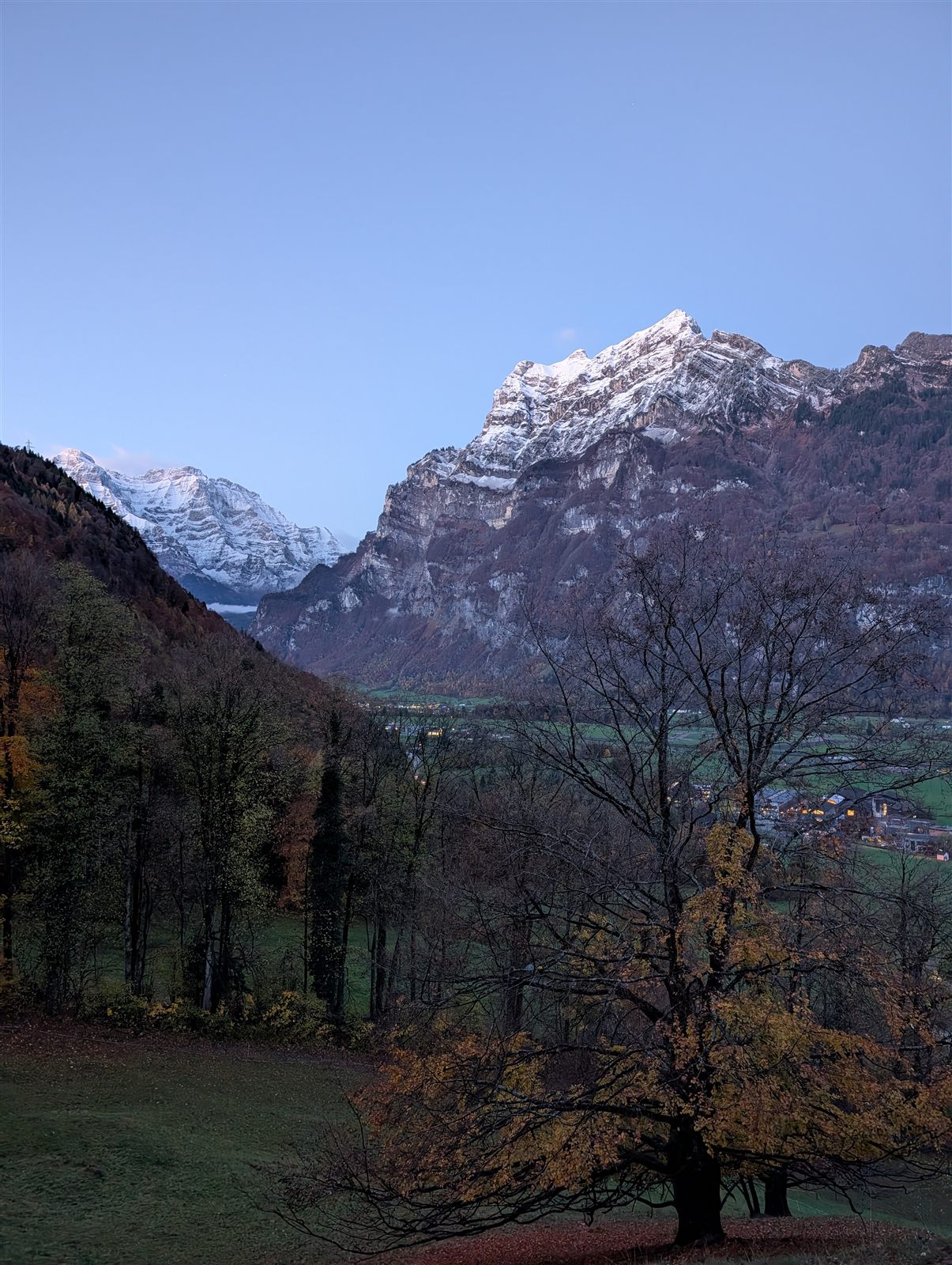 Snow-capped peaks above the Linth valley at dusk, seen from Mollis, with autumn trees in the foreground and village lights below.