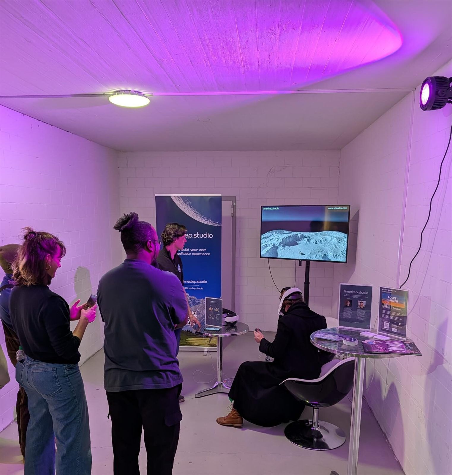 Visitors in a purple-lit room at the Swiss Space Museum; one sits in a headset while a moon landscape shows on screen.
